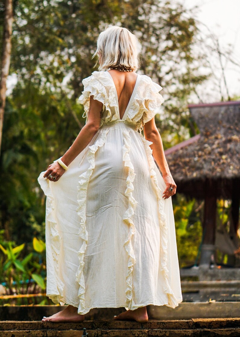 Puede incluir: Vestido largo color crema con volantes y escote en V pronunciado. El vestido est&aacute; hecho de una tela ligera y tiene una silueta fluida. La modelo est&aacute; de pie sobre escalones de piedra en un jard&iacute;n.