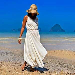 May include: A woman wearing a white maxi dress and a wide-brimmed straw hat walks along a sandy beach with blue water and islands in the background.