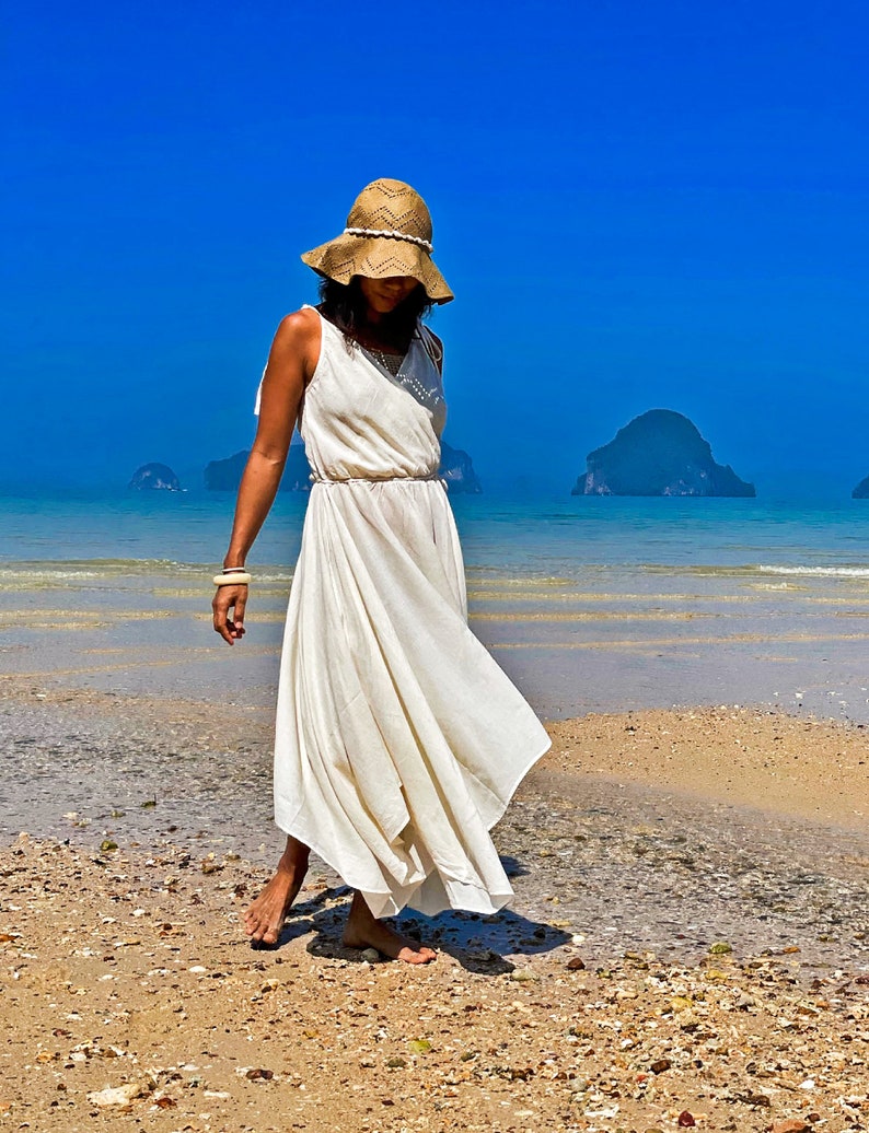 May include: A woman wearing a white maxi dress and a wide-brimmed straw hat walks along a sandy beach with blue water and islands in the background.