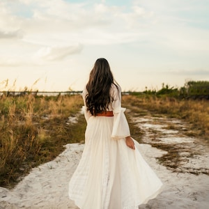 May include: A woman in a white maxi dress with a brown belt walks down a dirt path. The dress has a lace pattern and the woman's long brown hair is flowing in the wind.