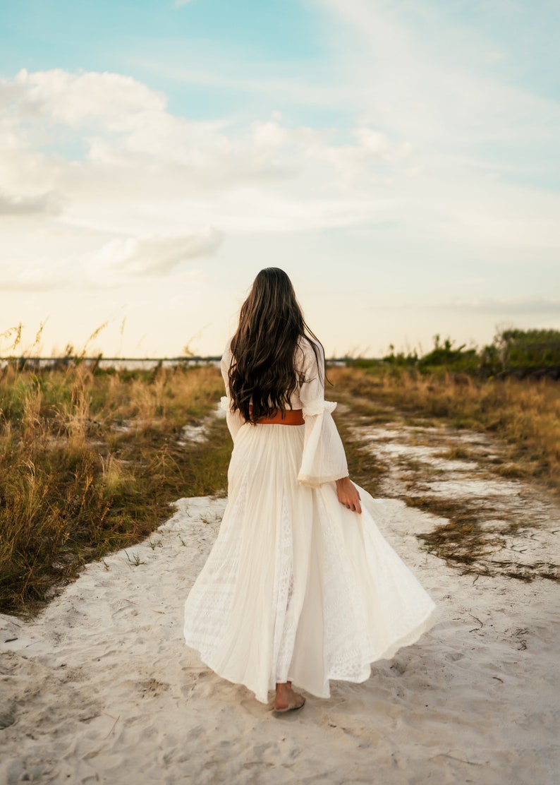May include: A woman in a white maxi dress with a brown belt walks down a dirt path. The dress has a lace pattern and the woman's long brown hair is flowing in the wind.