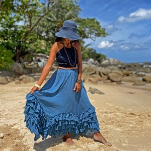 May include: A woman wearing a blue denim skirt with multiple ruffles, a black halter top, and a wide-brimmed blue hat. She is standing on a sandy beach with rocks and trees in the background.