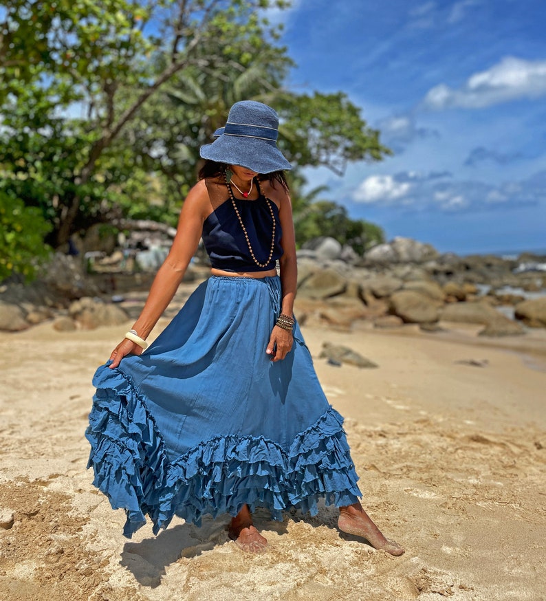 May include: A woman wearing a blue denim skirt with multiple ruffles, a black halter top, and a wide-brimmed blue hat. She is standing on a sandy beach with rocks and trees in the background.