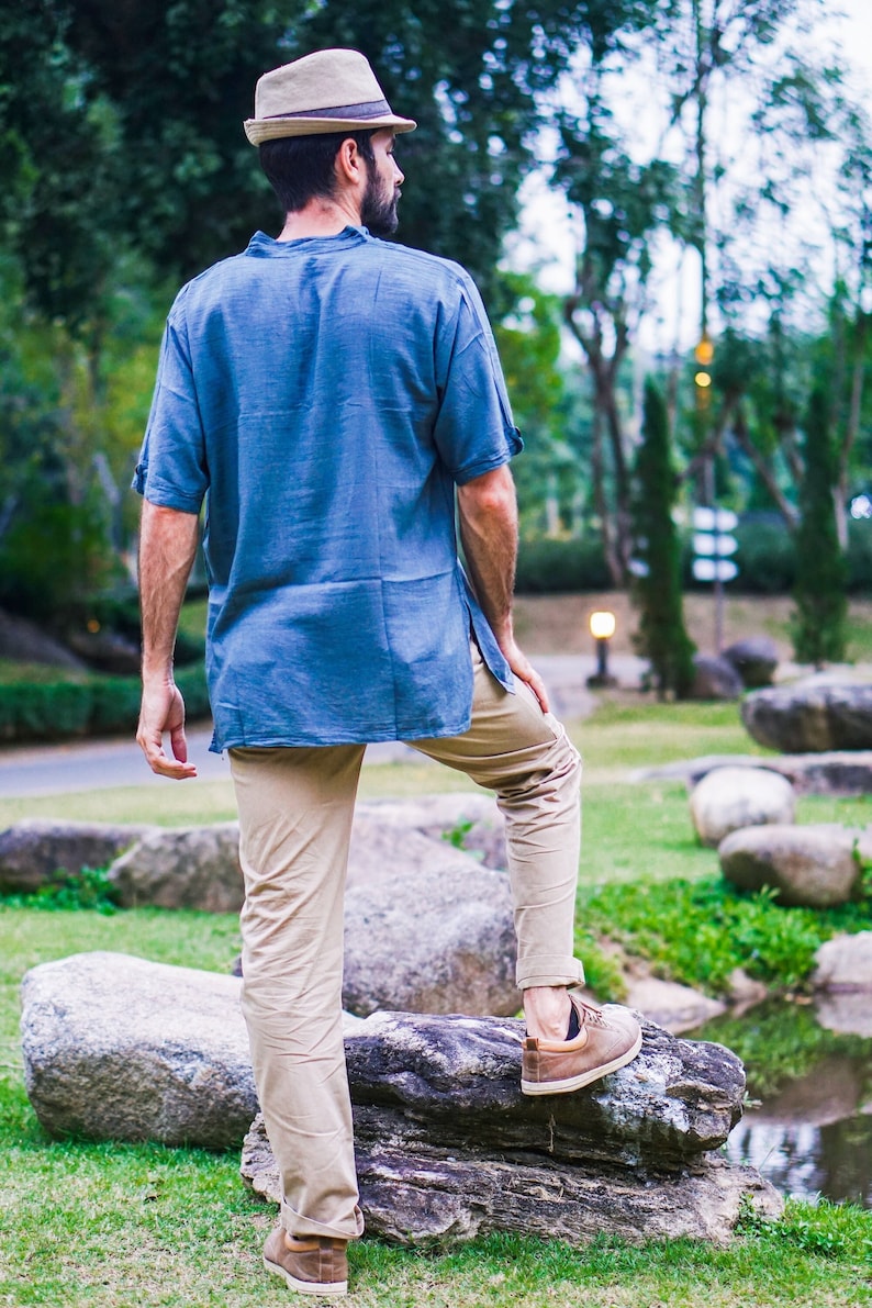 May include: A man wearing a light blue shirt, beige pants, and a brown fedora hat stands on a rock in a park setting. He is looking away from the camera.