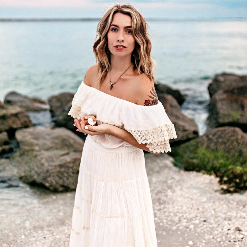 Puede incluir: Una persona con un vestido blanco con hombros descubiertos y ribete de encaje, de pie en una playa. Sostiene un peque&ntilde;o objeto blanco en sus manos. El fondo muestra rocas y el mar.