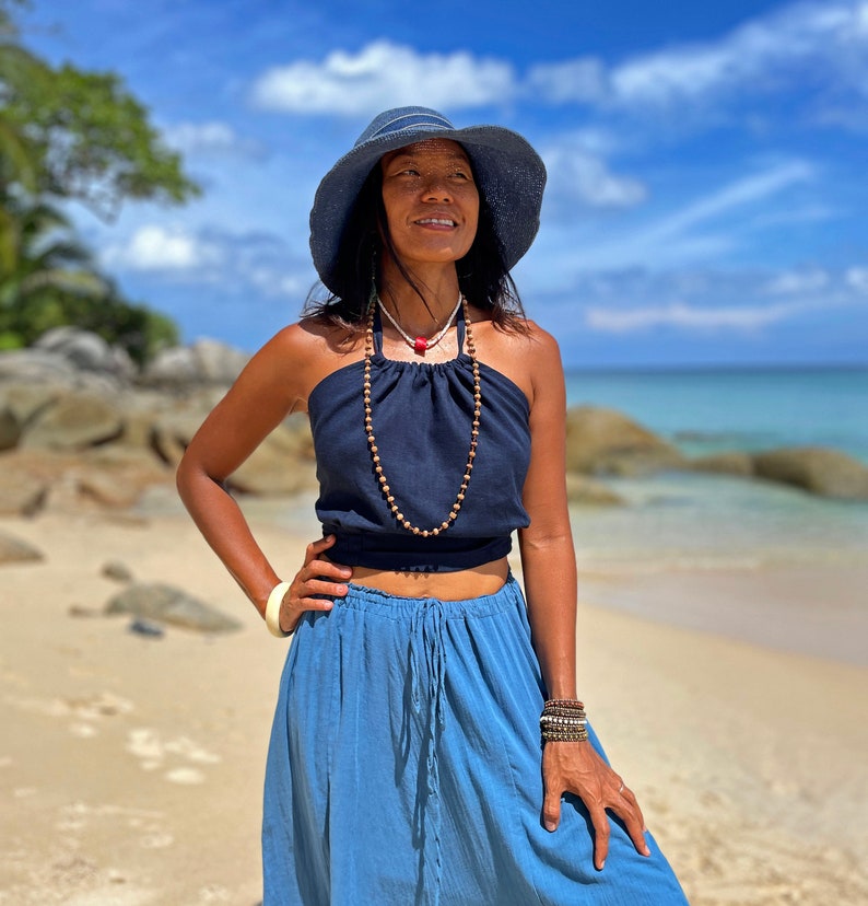 May include: A woman wearing a blue halter top with a brown beaded necklace, a blue skirt, and a blue sun hat. She is standing on a beach with a blue sky and white clouds in the background.