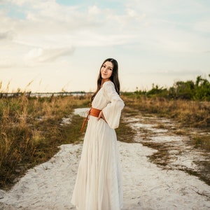May include: A woman in a white maxi dress with a brown leather belt stands on a sandy path. She is smiling and looking at the camera.