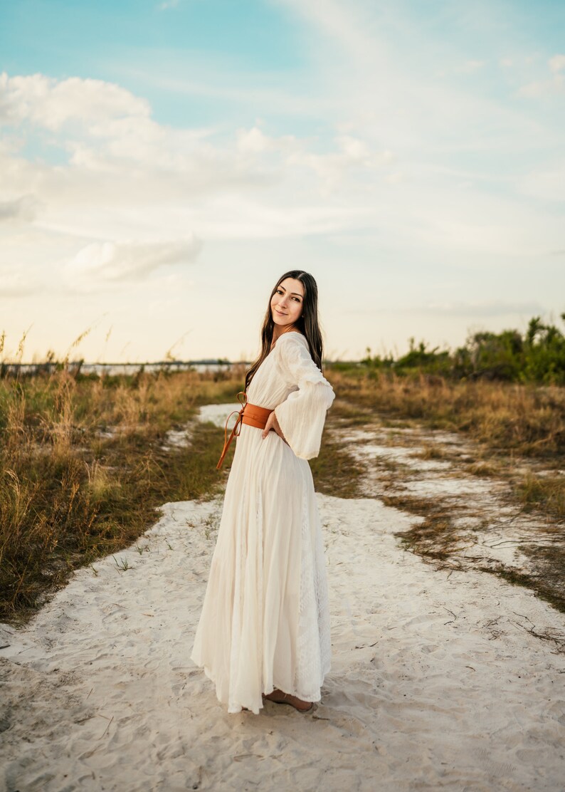 May include: A woman in a white maxi dress with a brown leather belt stands on a sandy path. She is smiling and looking at the camera.