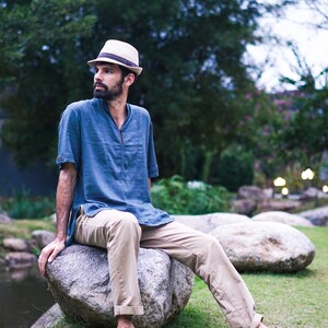 May include: A man wearing a blue shirt, khaki pants, and a straw hat sits on a large rock. He is looking off to the side.