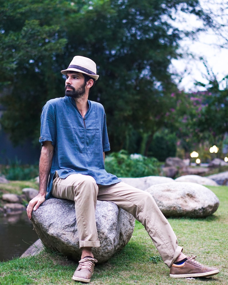 May include: A man wearing a blue shirt, khaki pants, and a straw hat sits on a large rock. He is looking off to the side.