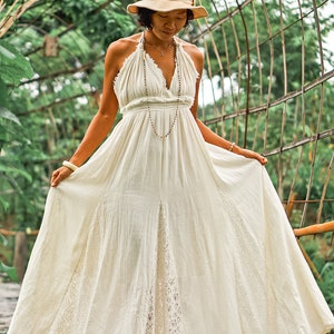 May include: A cream-colored halter maxi dress with a flowing skirt and a V-neckline. The dress features a lace panel on the skirt and is paired with a beige hat. A long beaded necklace and bracelets complete the look.