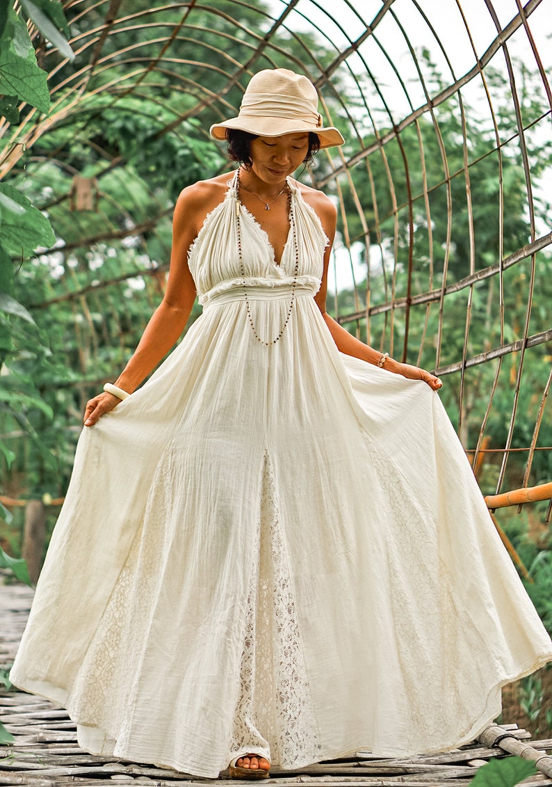 May include: A cream-colored halter maxi dress with a flowing skirt and a V-neckline. The dress features a lace panel on the skirt and is paired with a beige hat. A long beaded necklace and bracelets complete the look.