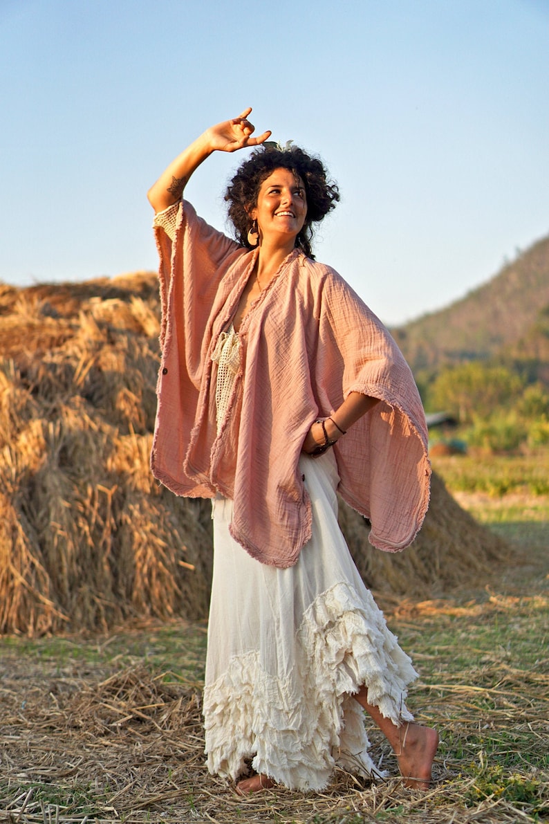 May include: A woman wearing a pink, loose-fitting,  boho-style top with a white, ruffled skirt. She is standing in a field of hay with a mountain in the background.