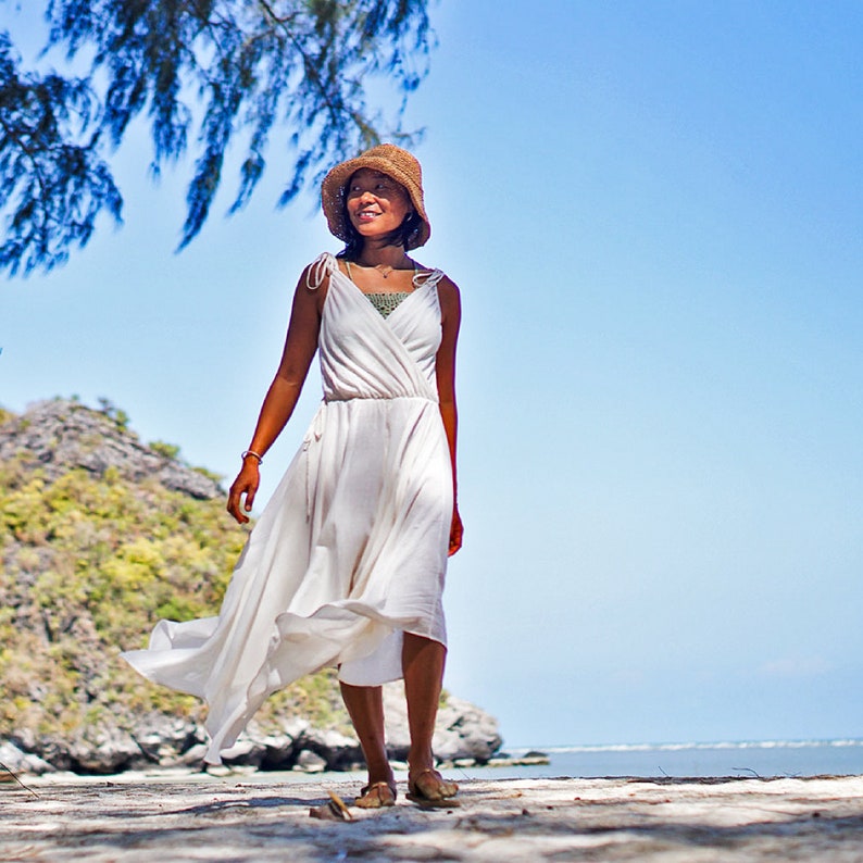May include: A woman wearing a white dress and a brown hat walks on a sandy beach with a rocky hill in the background.