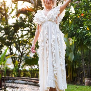 Puede incluir: Un vestido largo blanco con volantes y cuello en V. El vestido tiene una silueta fluida y se usa al aire libre. La imagen est&aacute; tomada con luz natural, con un fondo borroso de vegetaci&oacute;n y una fuente de agua.