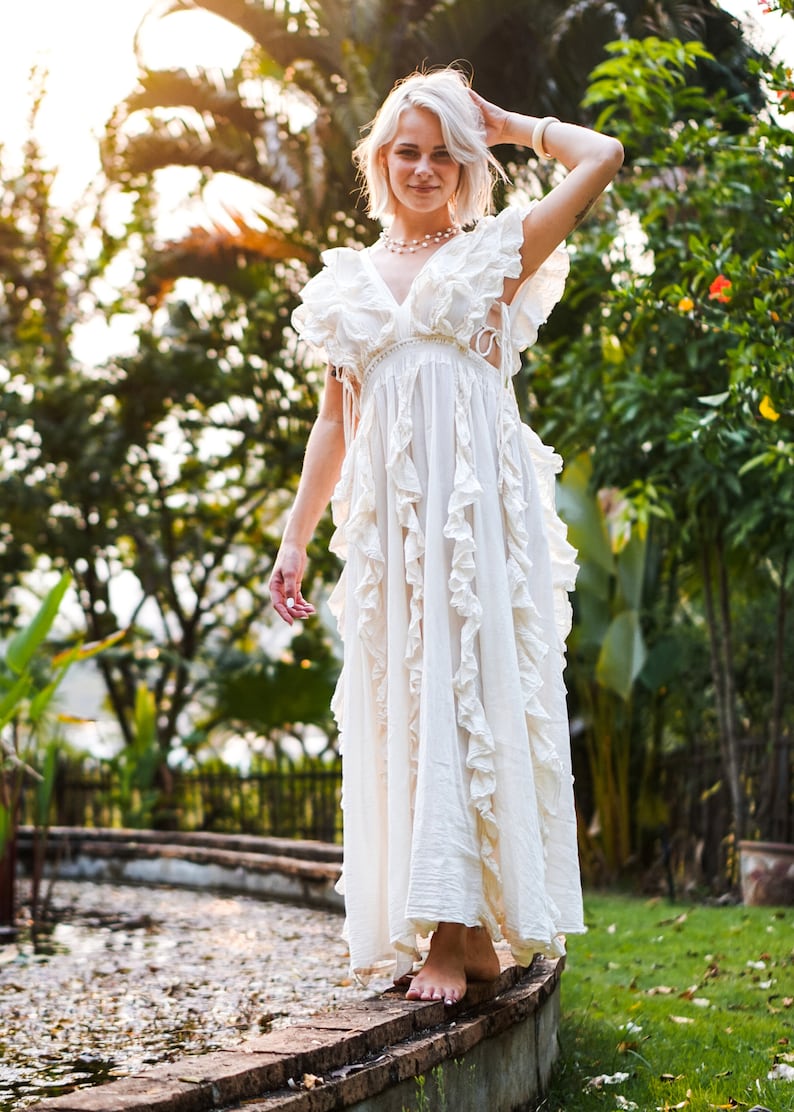 Puede incluir: Un vestido largo blanco con volantes y cuello en V. El vestido tiene una silueta fluida y se usa al aire libre. La imagen est&aacute; tomada con luz natural, con un fondo borroso de vegetaci&oacute;n y una fuente de agua.