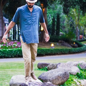 May include: A man wearing a blue shirt, khaki pants, and a straw hat walks across a path of large rocks in a park.