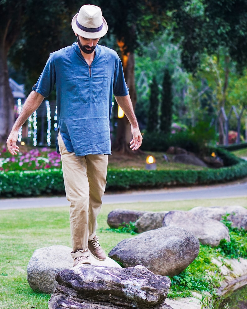 May include: A man wearing a blue shirt, khaki pants, and a straw hat walks across a path of large rocks in a park.