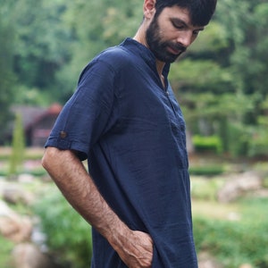 May include: A man wearing a dark blue short-sleeved shirt with a mandarin collar and a button closure. He is standing in a green grassy area with trees in the background.