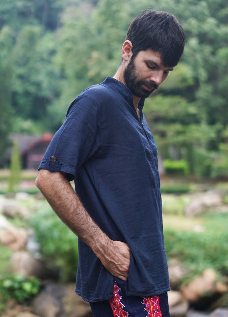 May include: A man wearing a dark blue short-sleeved shirt with a mandarin collar and a button closure. He is standing in a green grassy area with trees in the background.