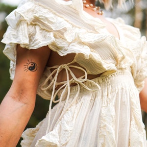 Puede incluir: Vestido color marfil con mangas y falda con volantes. El vestido presenta un escote en V y un detalle de espalda atada. Tambi&eacute;n se ven un collar de perlas y un tatuaje de un sol y el s&iacute;mbolo del yin-yang.