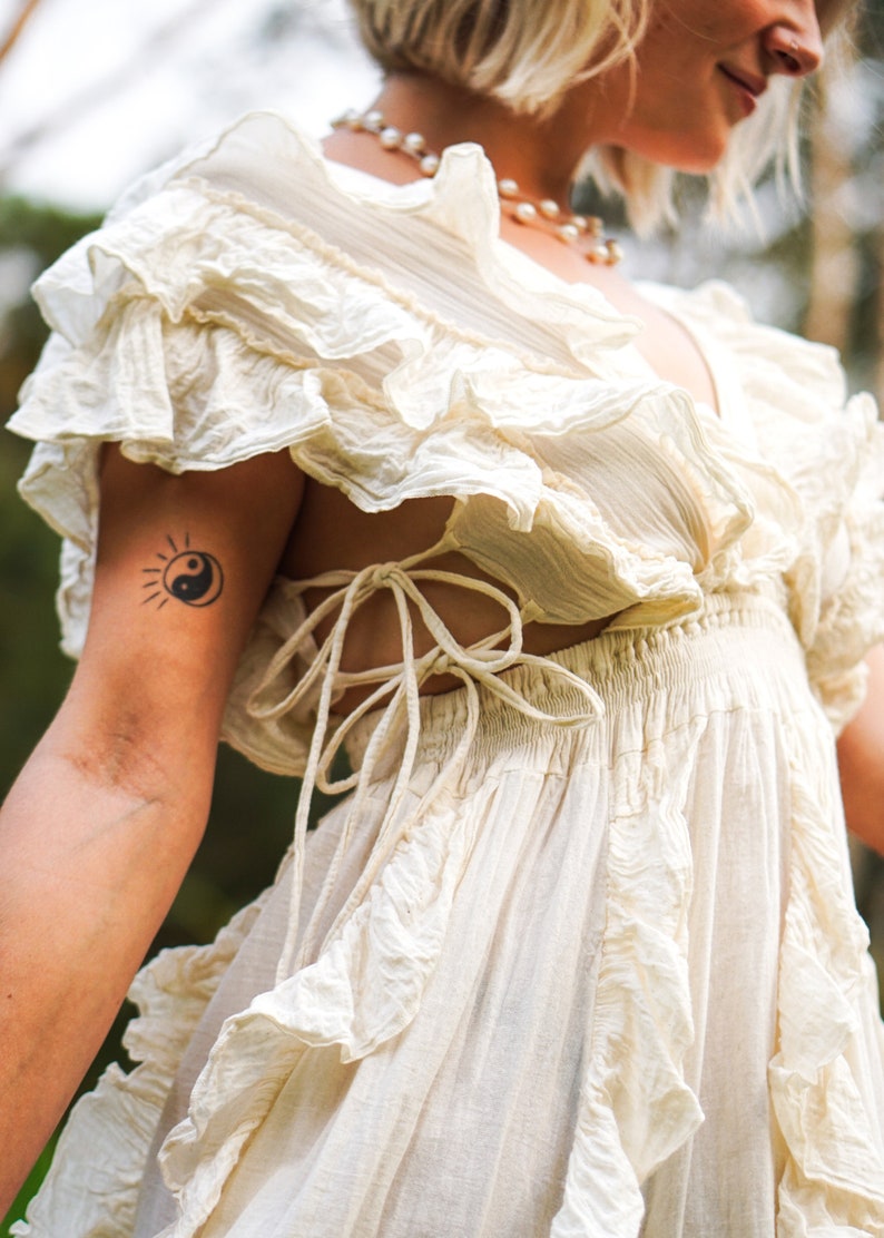 Puede incluir: Vestido color marfil con mangas y falda con volantes. El vestido presenta un escote en V y un detalle de espalda atada. Tambi&eacute;n se ven un collar de perlas y un tatuaje de un sol y el s&iacute;mbolo del yin-yang.