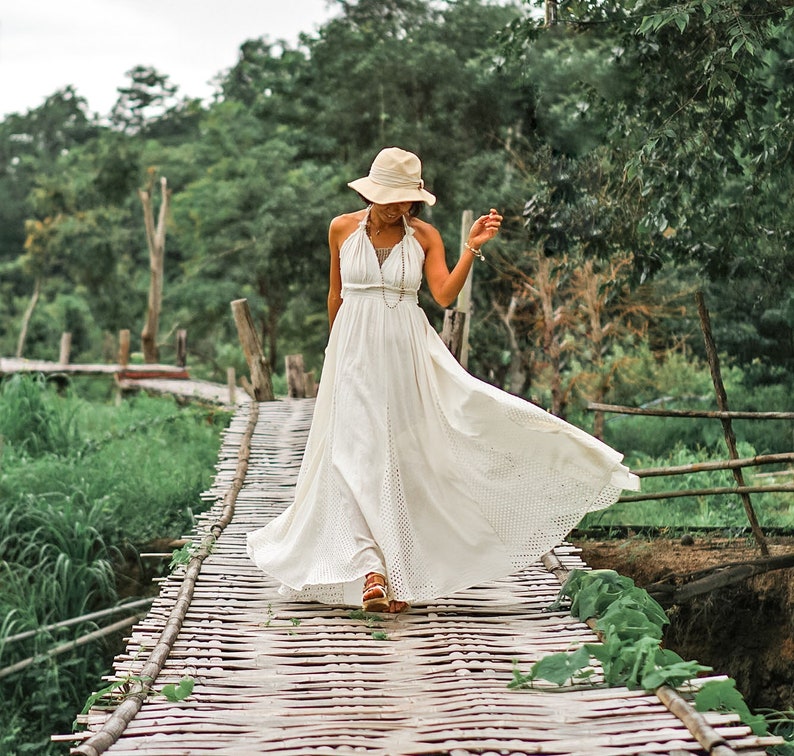 May include: A woman wearing a white halter maxi dress and a beige hat walks on a bamboo bridge. The dress has a flowing skirt with eyelet detailing. The background features lush green foliage and a wooden bridge.