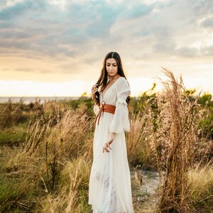 May include: A woman in a white maxi dress with a brown leather belt stands in a field of tall grass. The sky is cloudy and the sun is setting.