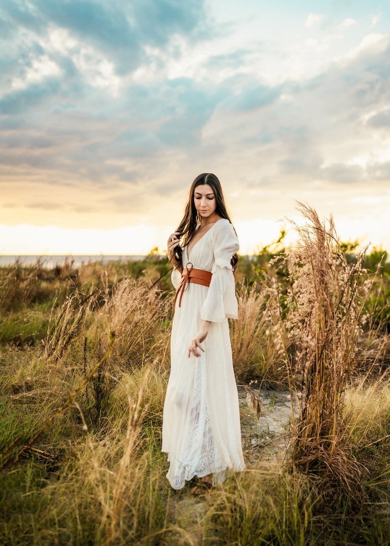 May include: A woman in a white maxi dress with a brown leather belt stands in a field of tall grass. The sky is cloudy and the sun is setting.