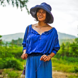 May include: A woman wearing a blue, loose-fitting top with a button closure and a matching blue skirt. She is wearing a blue sun hat and a necklace.