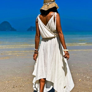 May include: A woman wearing a white maxi dress with a straw hat walks on a sandy beach. The ocean is in the background.