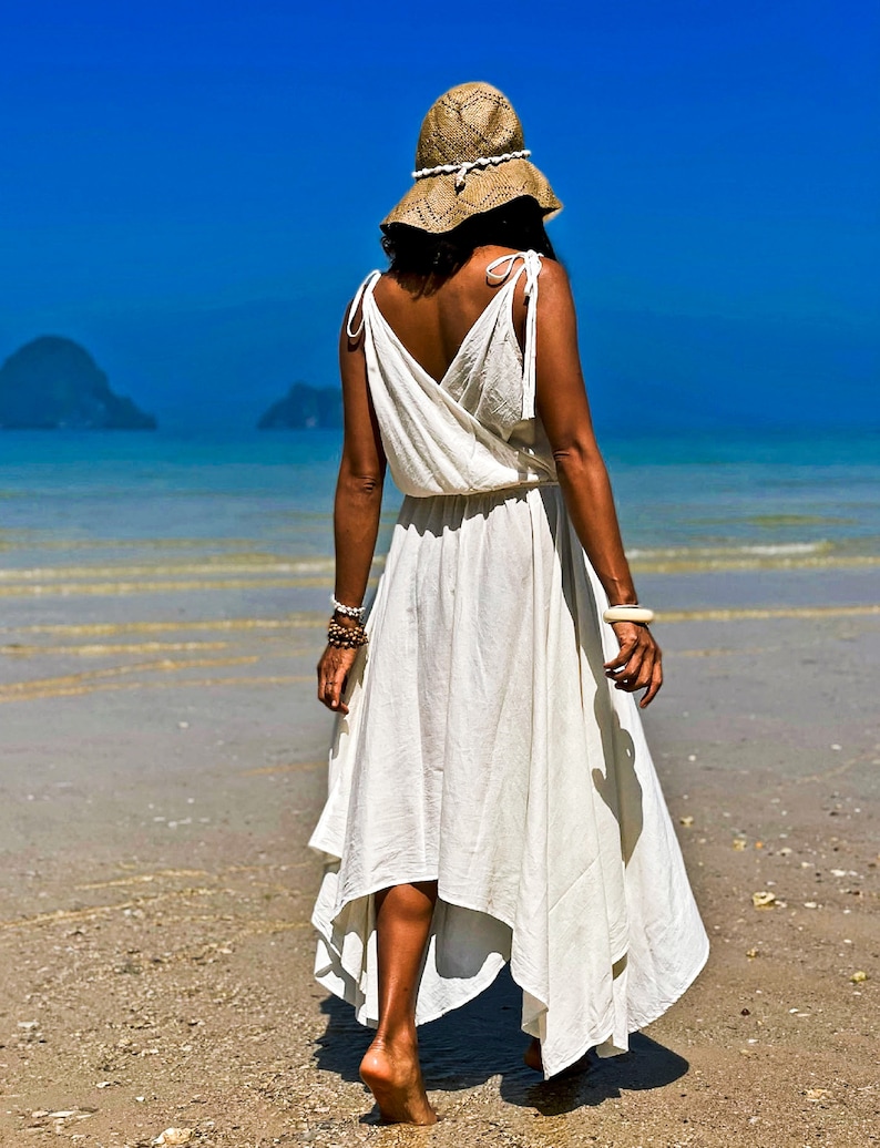 May include: A woman wearing a white maxi dress with a straw hat walks on a sandy beach. The ocean is in the background.
