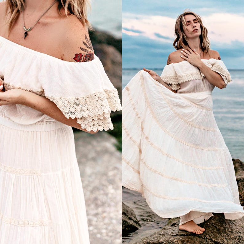 Puede incluir: Vestido blanco con hombros descubiertos y encaje en las mangas y la falda. El vestido tiene una falda fluida y escalonada y se usa al aire libre cerca del agua. La tela es ligera.