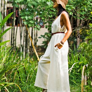 May include: A woman wearing a white linen jumpsuit with a brown belt and a straw hat. She is standing in a garden with green plants.