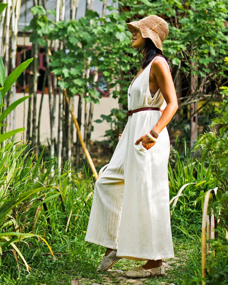May include: A woman wearing a white linen jumpsuit with a brown belt and a straw hat. She is standing in a garden with green plants.