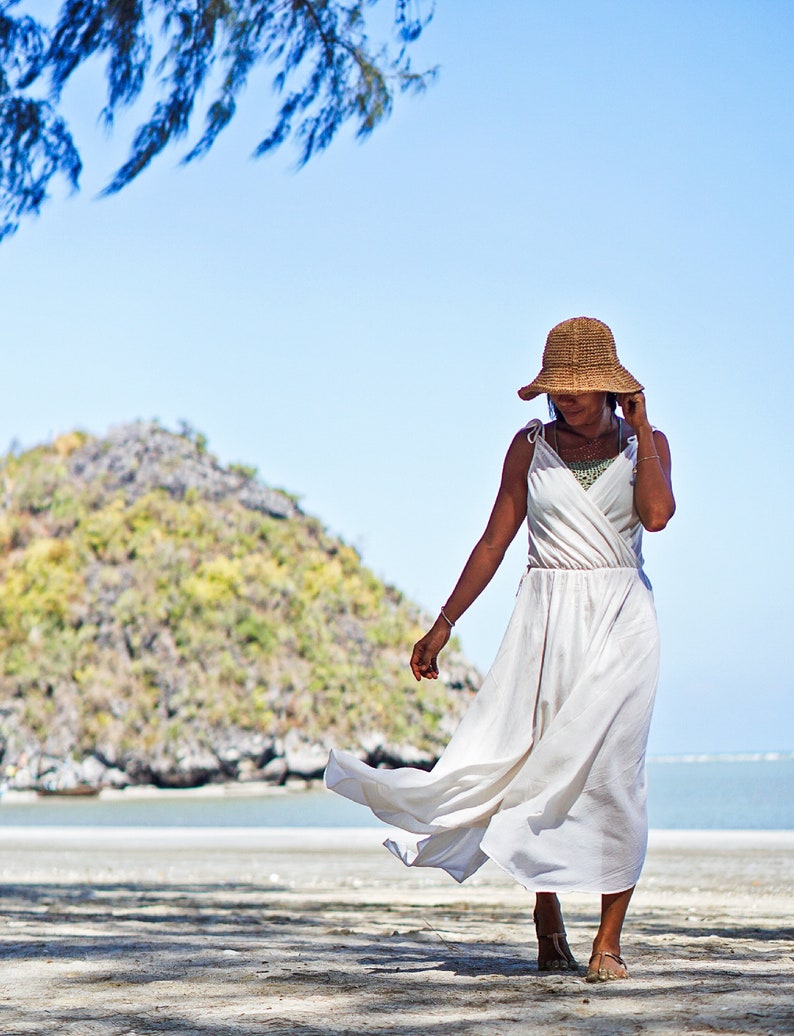 May include: A woman in a white maxi dress and a wide-brimmed straw hat walks on a sandy beach. The beach is bordered by a body of water and a rocky hill covered in vegetation.