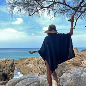 K&ouml;nnte beinhalten: Eine Frau in einem schwarzen Kaftan und einem Strohhut mit schwarzer Schleife steht auf einer felsigen Klippe mit Blick auf das Meer.