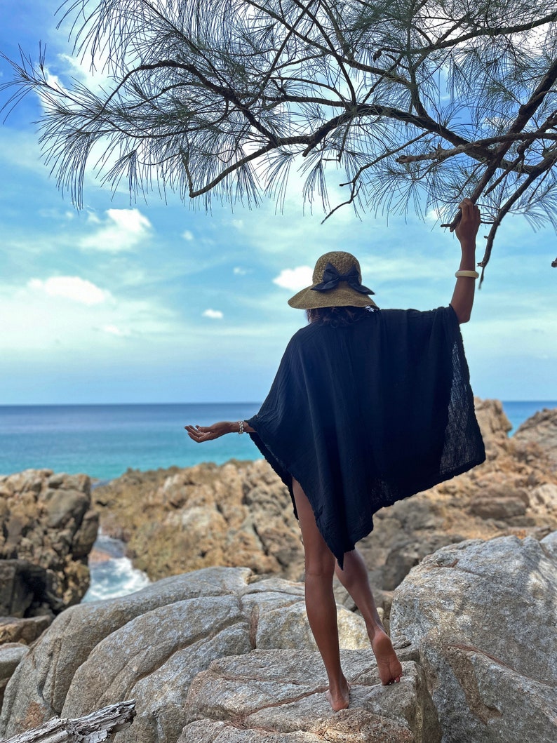 K&ouml;nnte beinhalten: Eine Frau in einem schwarzen Kaftan und einem Strohhut mit schwarzer Schleife steht auf einer felsigen Klippe mit Blick auf das Meer.