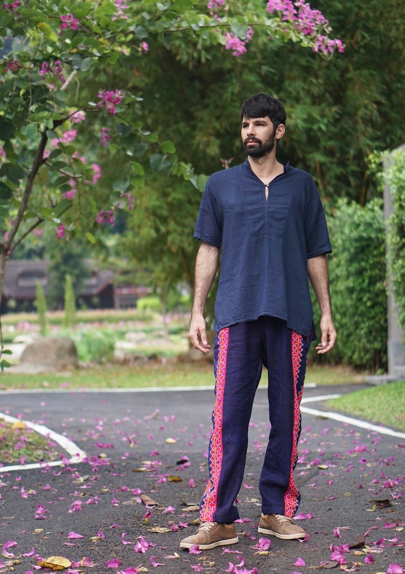 May include: A man wearing a dark blue shirt and dark blue pants with colorful embroidered details. He is standing on a paved path with pink flower petals scattered on the ground.