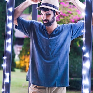 May include: A man wearing a blue shirt and khaki pants stands in a doorway with string lights. He is wearing a beige hat with a black band.