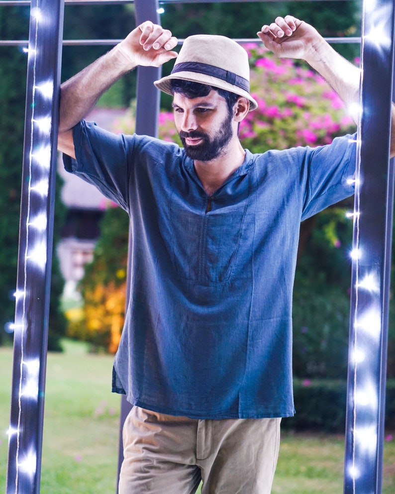May include: A man wearing a blue shirt and khaki pants stands in a doorway with string lights. He is wearing a beige hat with a black band.