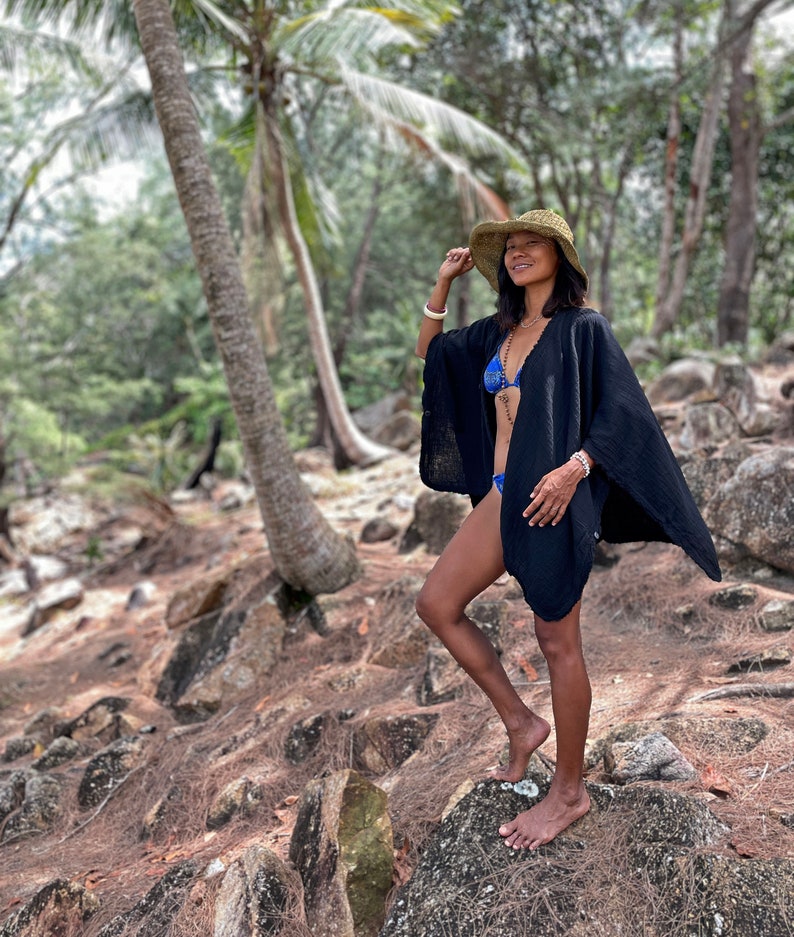 K&ouml;nnte beinhalten: Eine Frau tr&auml;gt einen schwarzen Leinen-Kimono mit einem blauen Bikini-Oberteil darunter. Sie steht auf einem Felsen in einer tropischen Waldlandschaft. Sie tr&auml;gt einen Strohhut und hat ihr Haar offen.
