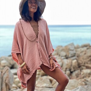 May include: A woman wearing a pink, loose-fitting, cotton cover-up with a brown straw hat. She is standing on a rocky beach with the ocean in the background.