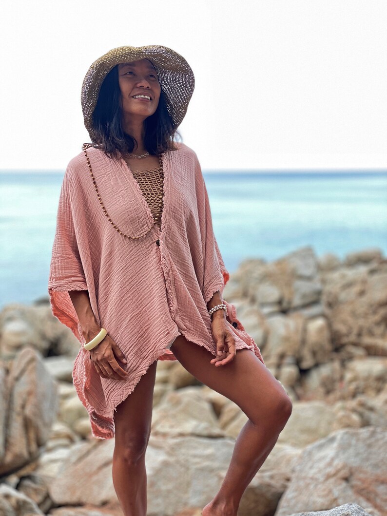 May include: A woman wearing a pink, loose-fitting, cotton cover-up with a brown straw hat. She is standing on a rocky beach with the ocean in the background.
