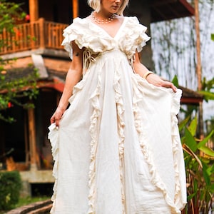 Puede incluir: Un vestido largo color crema con volantes y escote en V. El vestido presenta mangas cortas con volantes y una falda fluida. La modelo est&aacute; de pie al aire libre, con un edificio de madera al fondo. El vestido parece estar hecho de una tela ligera.