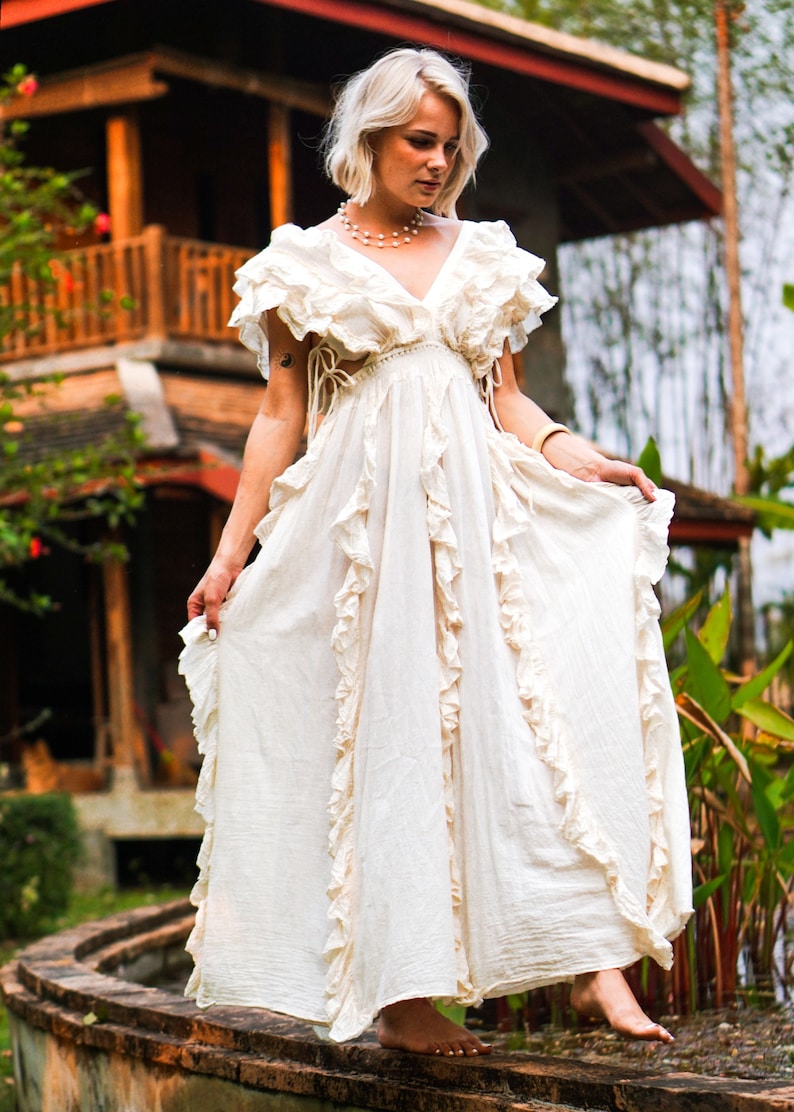 Puede incluir: Un vestido largo color crema con volantes y escote en V. El vestido presenta mangas cortas con volantes y una falda fluida. La modelo est&aacute; de pie al aire libre, con un edificio de madera al fondo. El vestido parece estar hecho de una tela ligera.