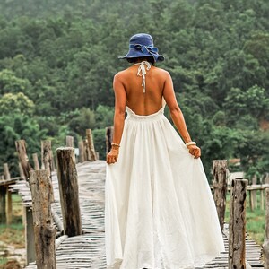 May include: A woman wearing a white halter dress and a blue hat with a bow, walking on a wooden bridge. The dress is long and flowing. The background features a lush green forest and a cloudy sky.