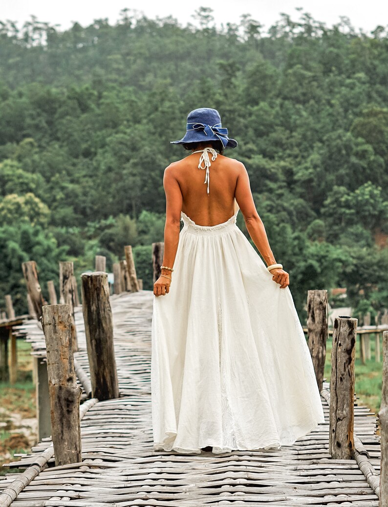 May include: A woman wearing a white halter dress and a blue hat with a bow, walking on a wooden bridge. The dress is long and flowing. The background features a lush green forest and a cloudy sky.