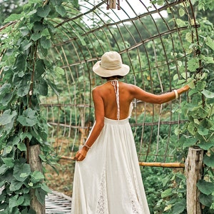 May include: A woman in a white halter dress and a wide-brimmed hat stands on a bamboo bridge, reaching towards a metal arch covered in green vines. The dress has lace detailing and a flowing skirt, creating a serene, natural aesthetic.
