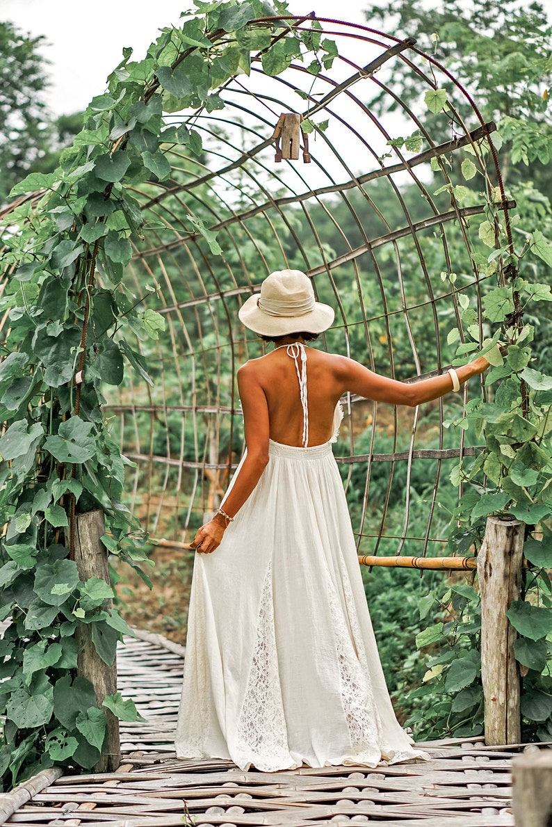 May include: A woman in a white halter dress and a wide-brimmed hat stands on a bamboo bridge, reaching towards a metal arch covered in green vines. The dress has lace detailing and a flowing skirt, creating a serene, natural aesthetic.